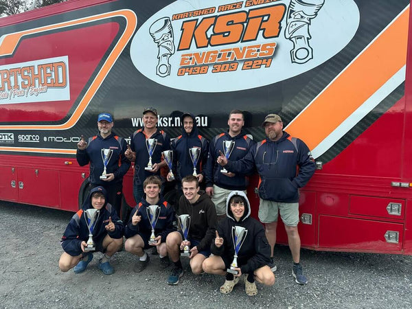 Group of people posing with trophies in front of a Kart Shed Racing Engines branded bus.
